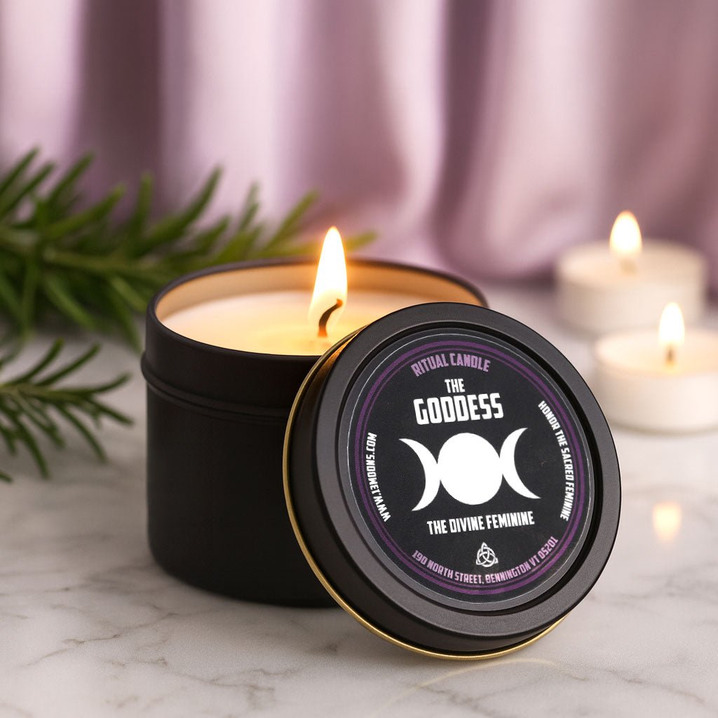Candle in a black tin labeled 'The Goddess' with a purple design, surrounded by other candles and greenery.